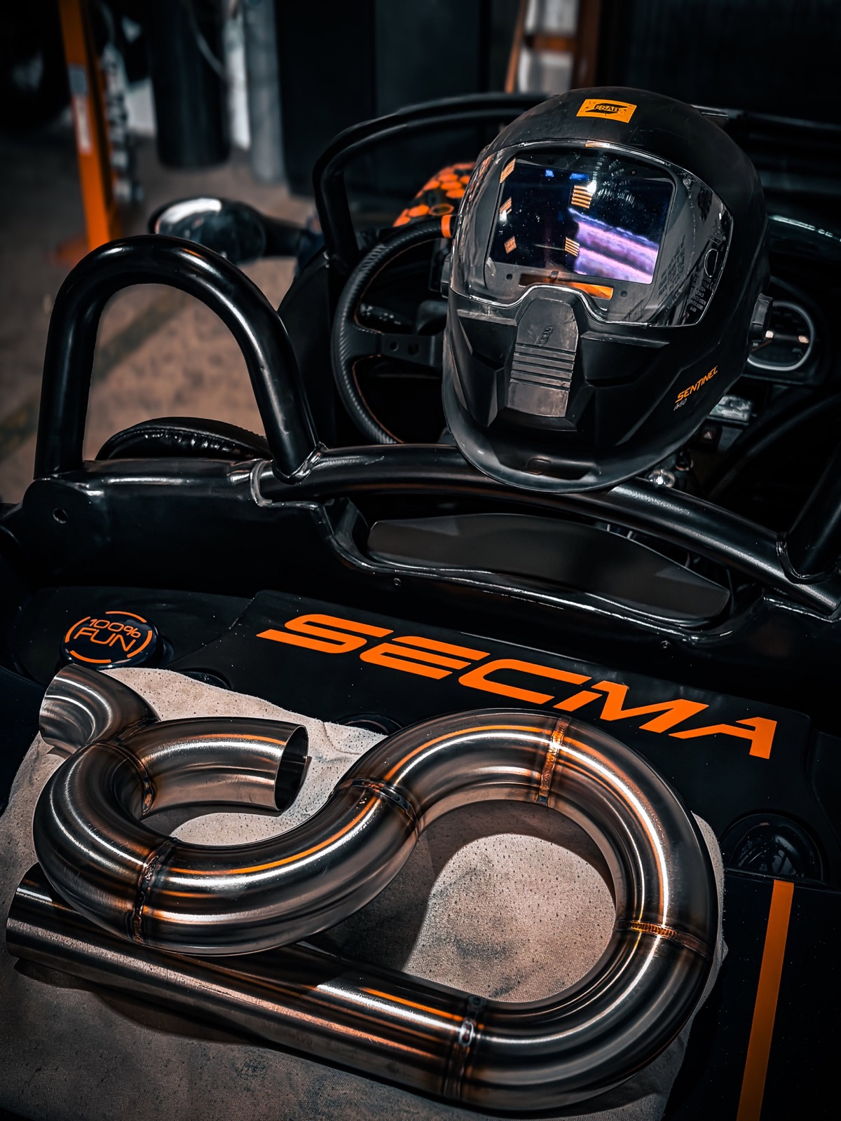 Ligne d'échappement inox cintrée posée sur un buggy Secma, casque de soudure en arrière-plan.